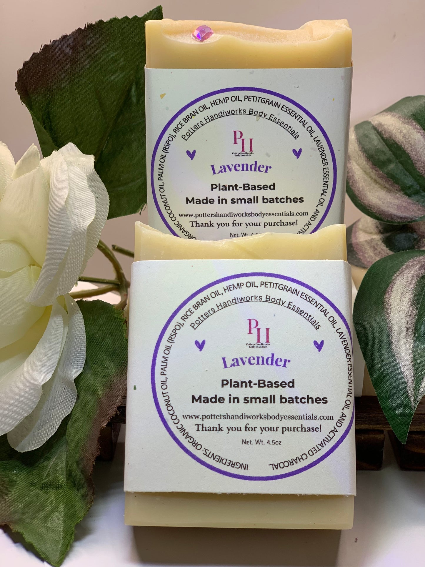 Two lavender soap bars with purple labels on a white surface with green leaves in the background.